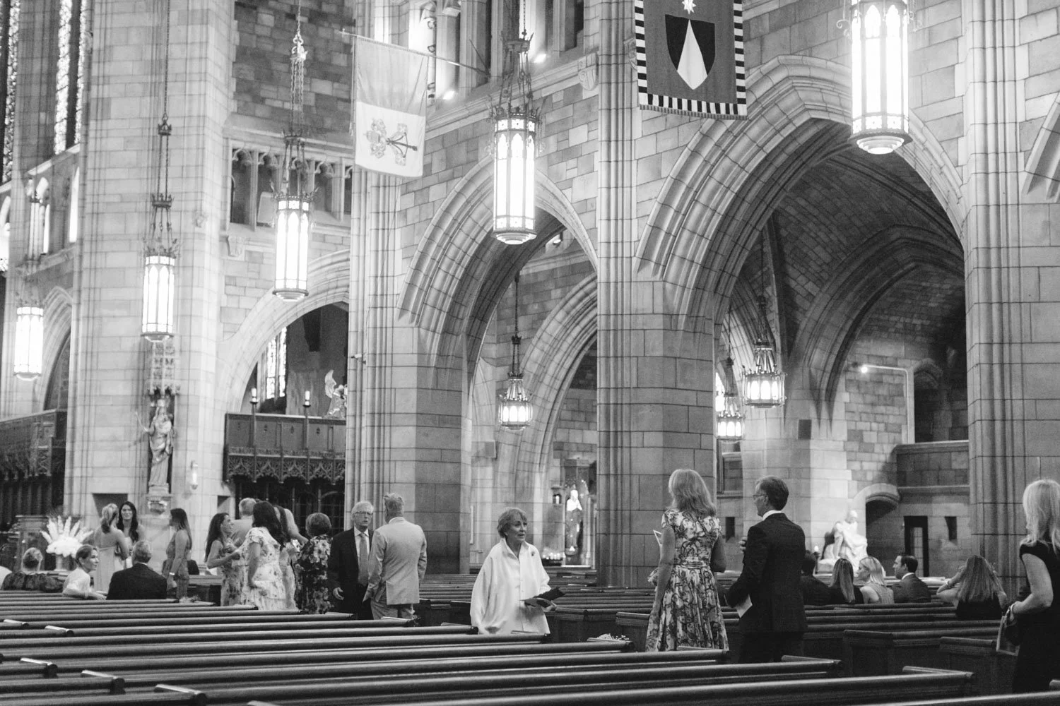 People gathered inside a large, stone church with high vaulted ceilings, stained glass windows, and hanging lantern lights, engaging in conversations.
