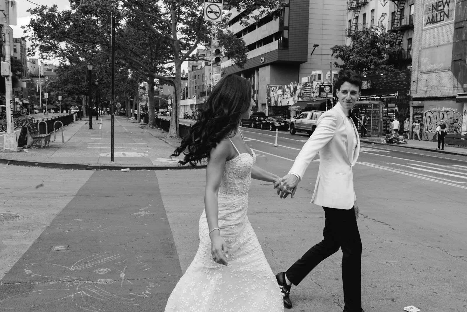 A couple walks hand in hand across a city street, with the woman in a wedding dress and the man in a white jacket.