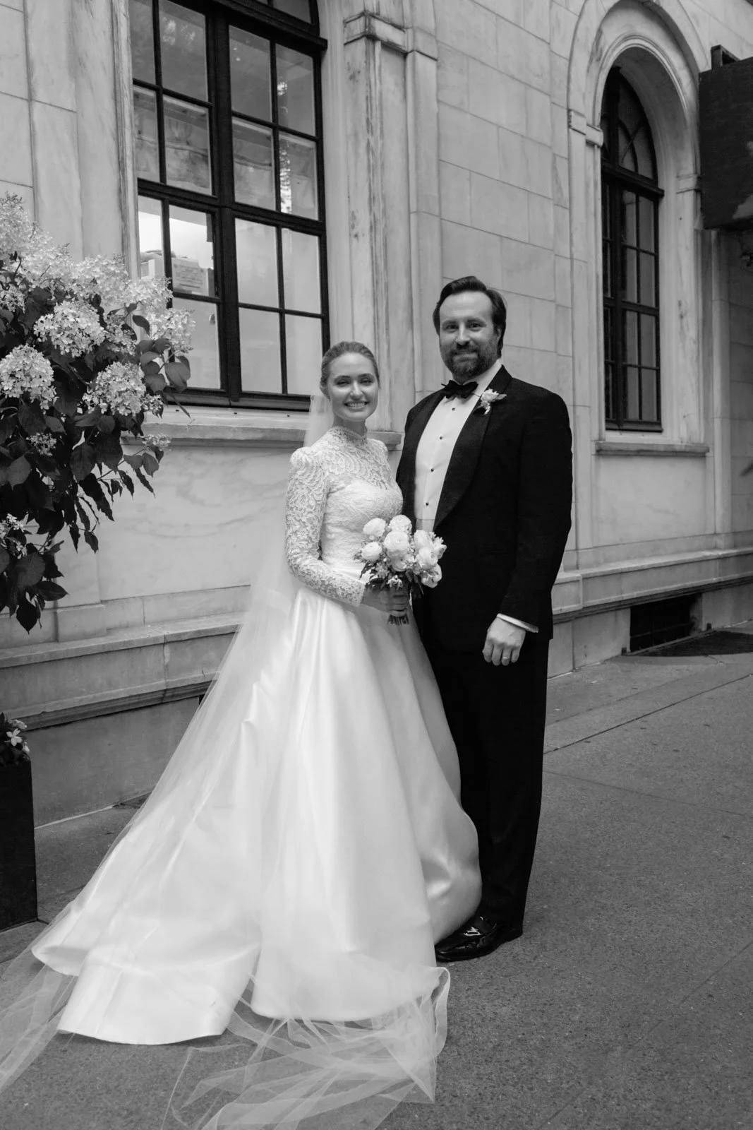 Black and white photograph of a bride and groom standing outside a building. The bride is holding a bouquet and is wearing a lace wedding gown with a long train. The groom is dressed in a tuxedo with a bow tie. They are smiling and standing close tog