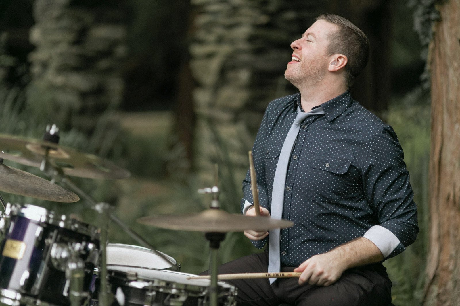 A man happily playing drums outdoors, wearing a dark blue polka dot shirt, a gray tie, and black pants.