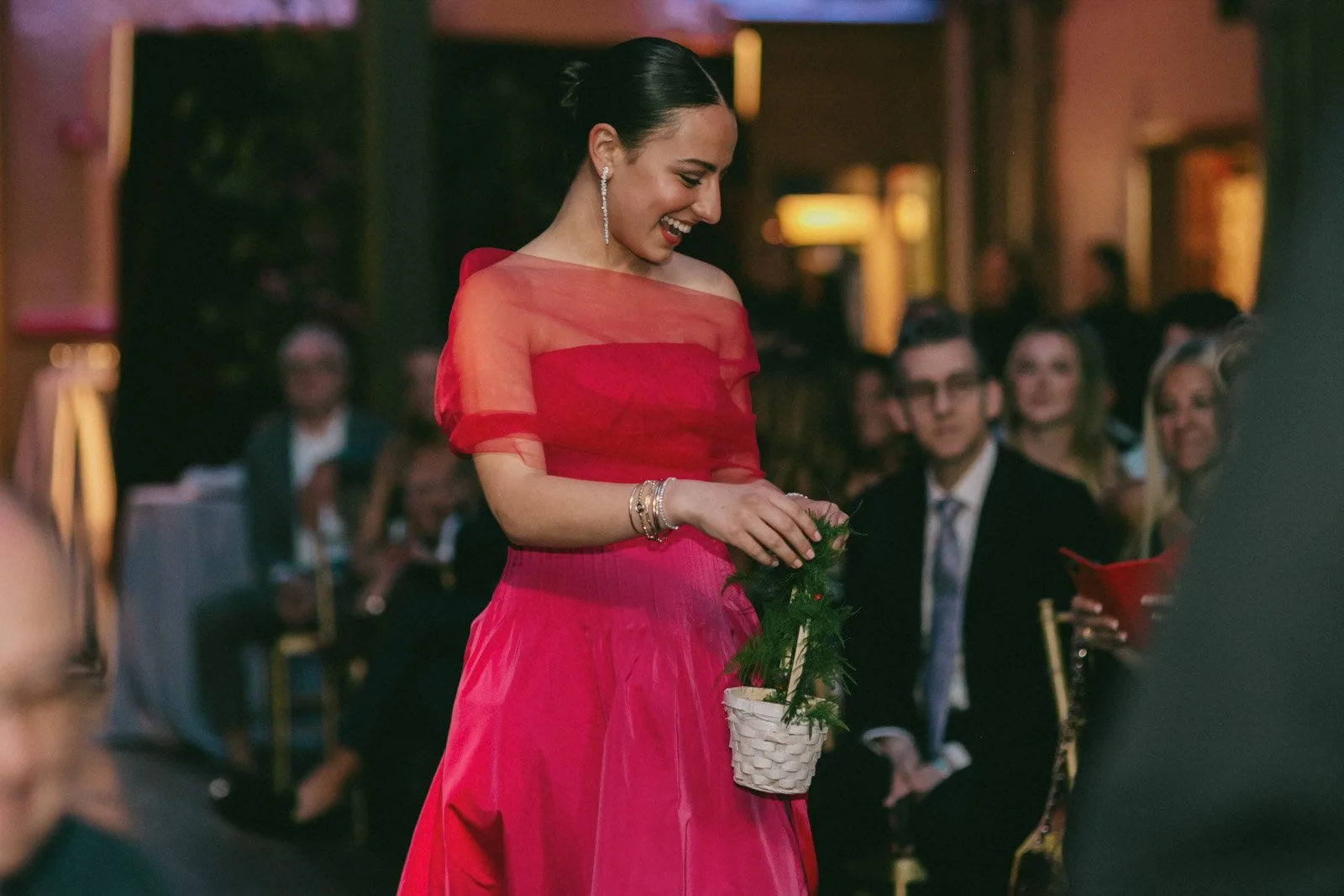 A woman in a bright pink dress is smiling and holding a small basket with greenery inside at an indoor event, with seated guests watching in the background.