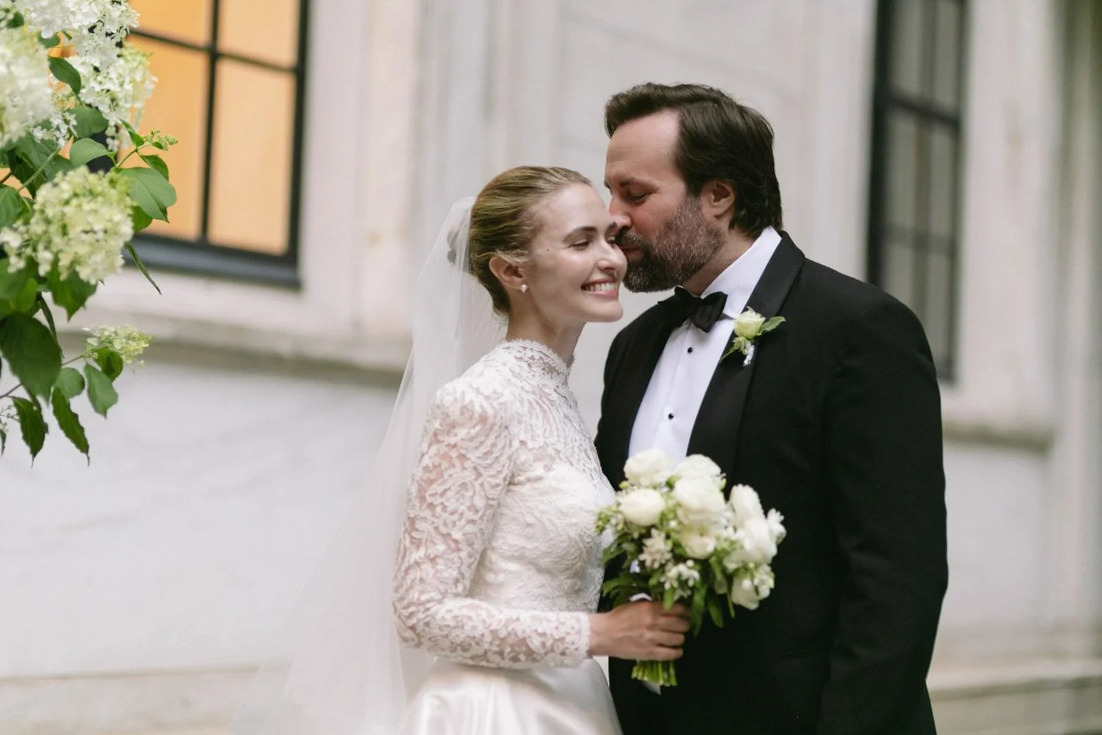 A bride and groom share a tender moment on their wedding day, smiling and close with the bride holding a bouquet of white flowers, dressed in a lace wedding gown, and the groom in a black tuxedo with a bow tie and boutonniere.