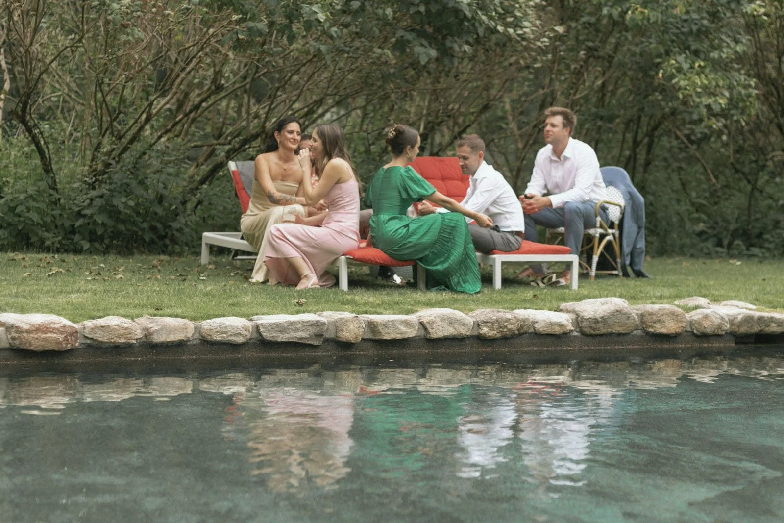 Group of six people sitting on outdoor benches on a grassy area near a water feature, engaging in conversations and enjoying a gathering.