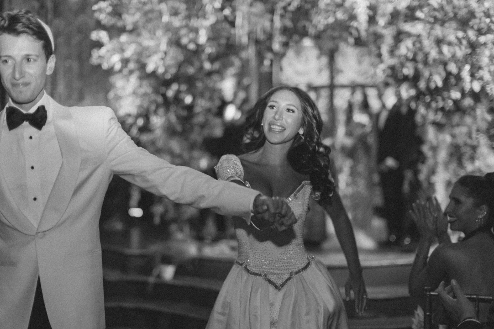 A black-and-white photo of a couple dancing at a formal event, with the woman smiling and the man holding her hand. There are other guests seated in the background, surrounded by trees.
