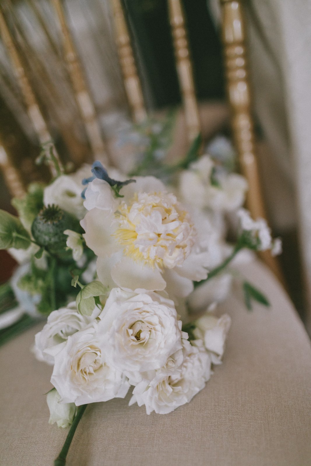 A bouquet of white flowers, possibly roses or peonies, resting on a beige surface. The background includes a decorative gold chair.