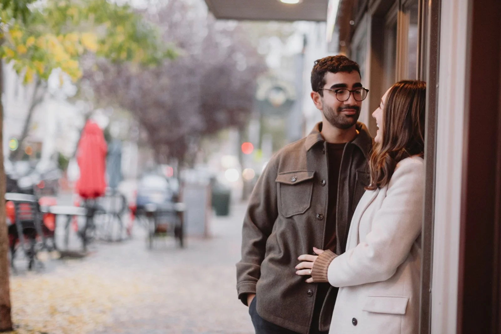 A man and woman stand close to each other on a sidewalk, engaging in a conversation. The man has dark hair, glasses, and a beard, wearing a dark jacket. The woman has long brown hair, wearing a white coat, and smiling. The background shows blurred tr