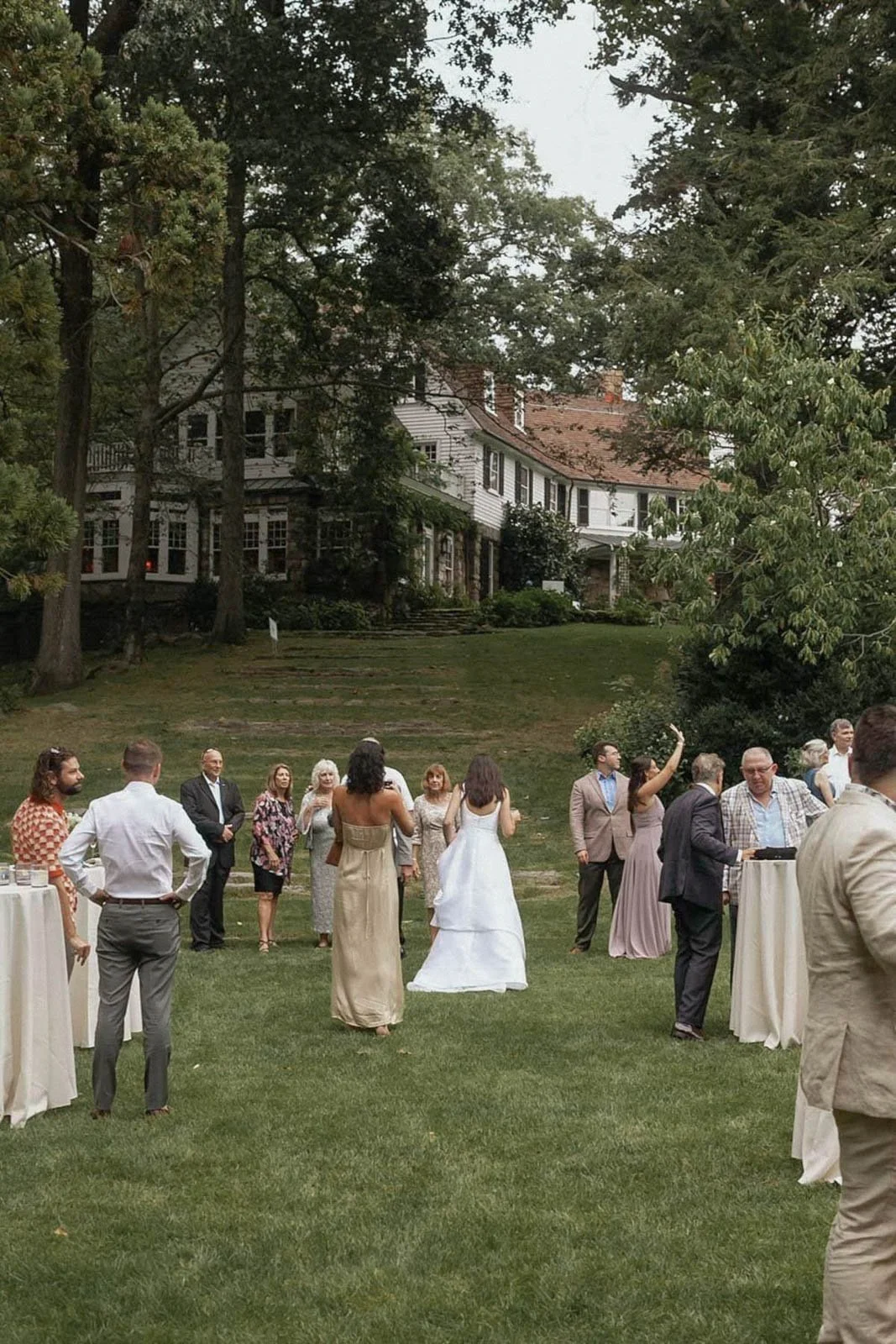 People gathered outdoors on a lawn, likely at a wedding or social event, with a large white house and trees in the background.