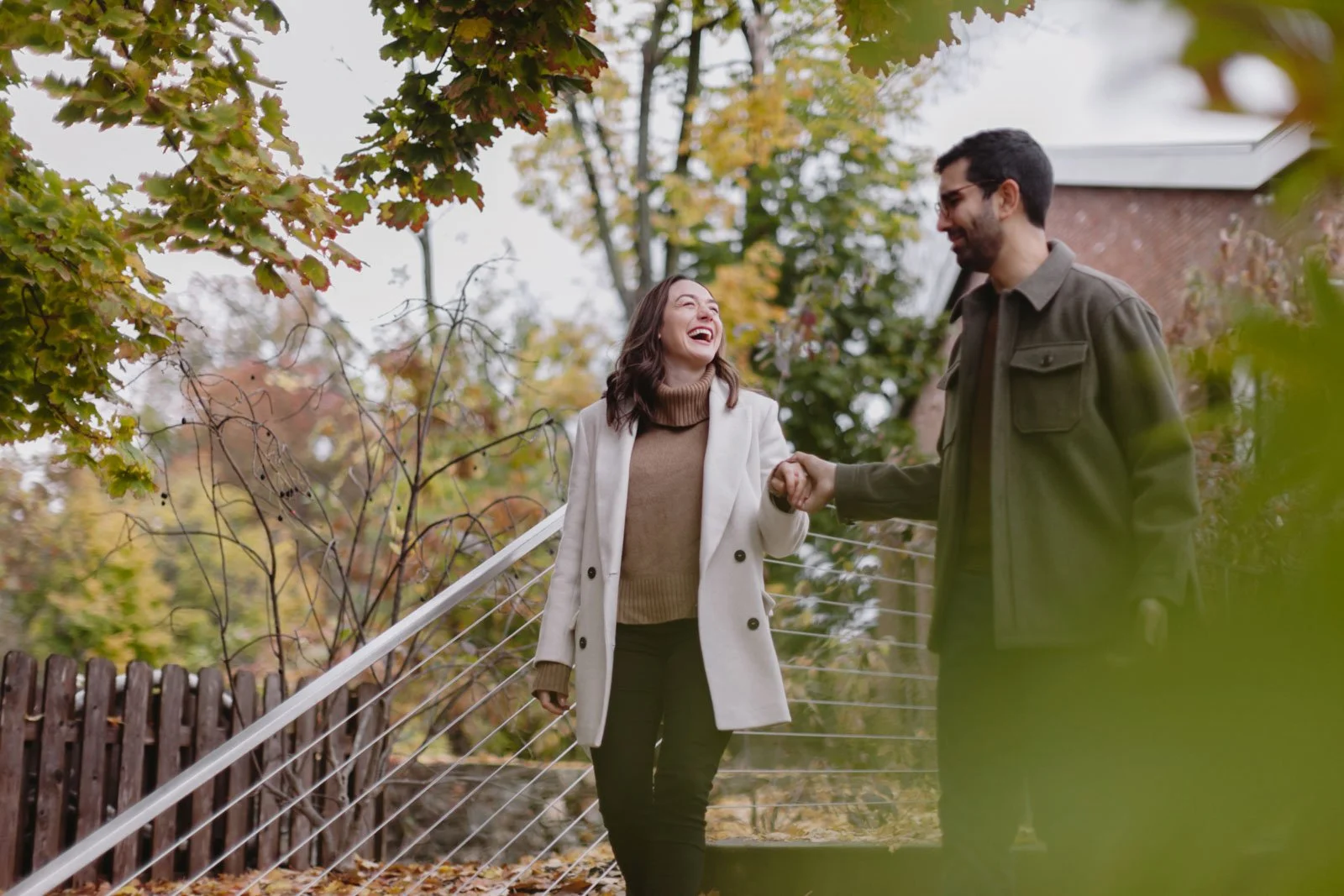 A man and woman holding hands and smiling in front of The roundhouse in Beacon, NY during fall, surrounded by trees with colorful leaves.