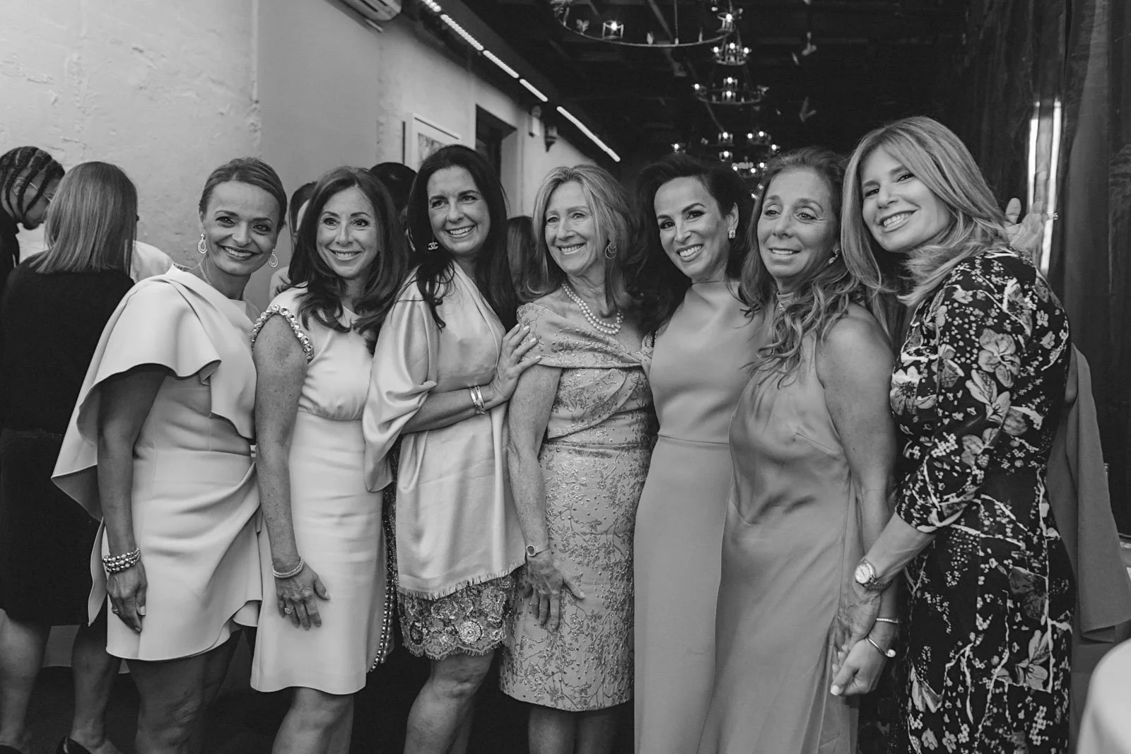 Ten women in formal dresses posing together at an indoor event, smiling for the camera.