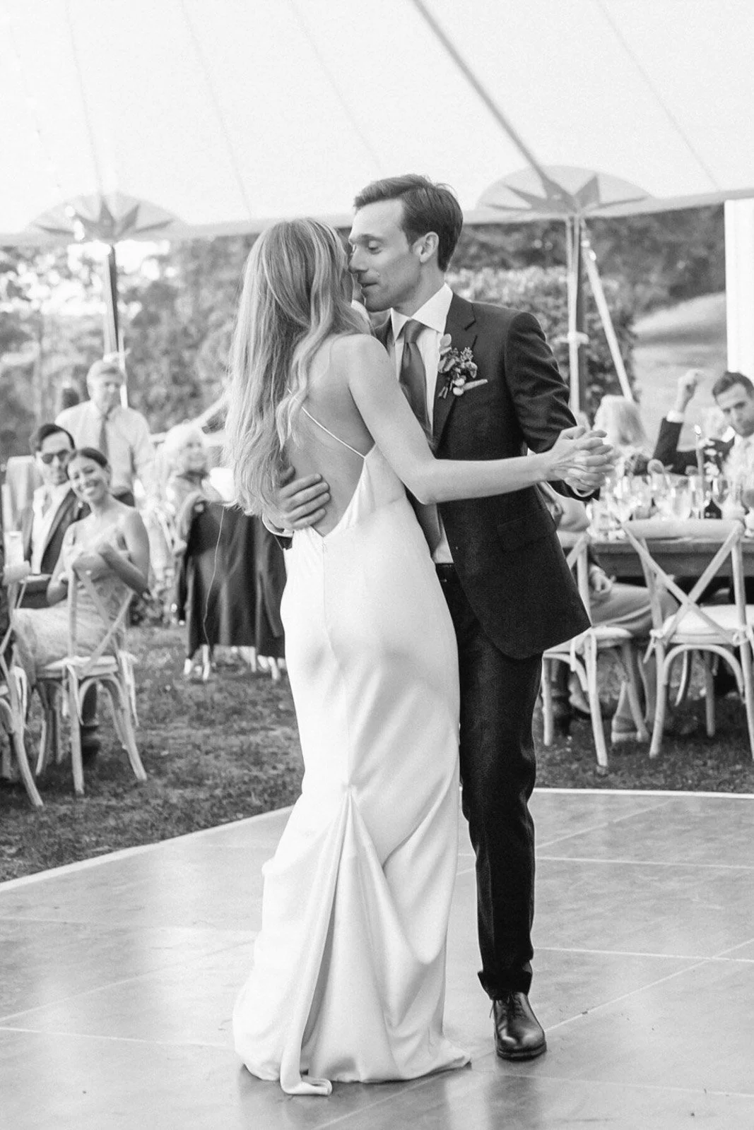 A black-and-white photo of a bride and groom dancing at their wedding reception, with guests seated at tables in the background under a tent.