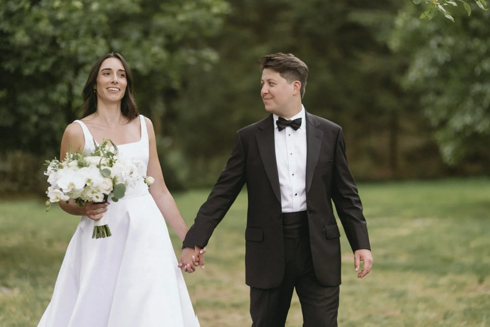 A bride and groom walk hand-in-hand outdoors, holding hands, with the bride holding a bouquet of flowers, and smiling at each other, surrounded by green trees.