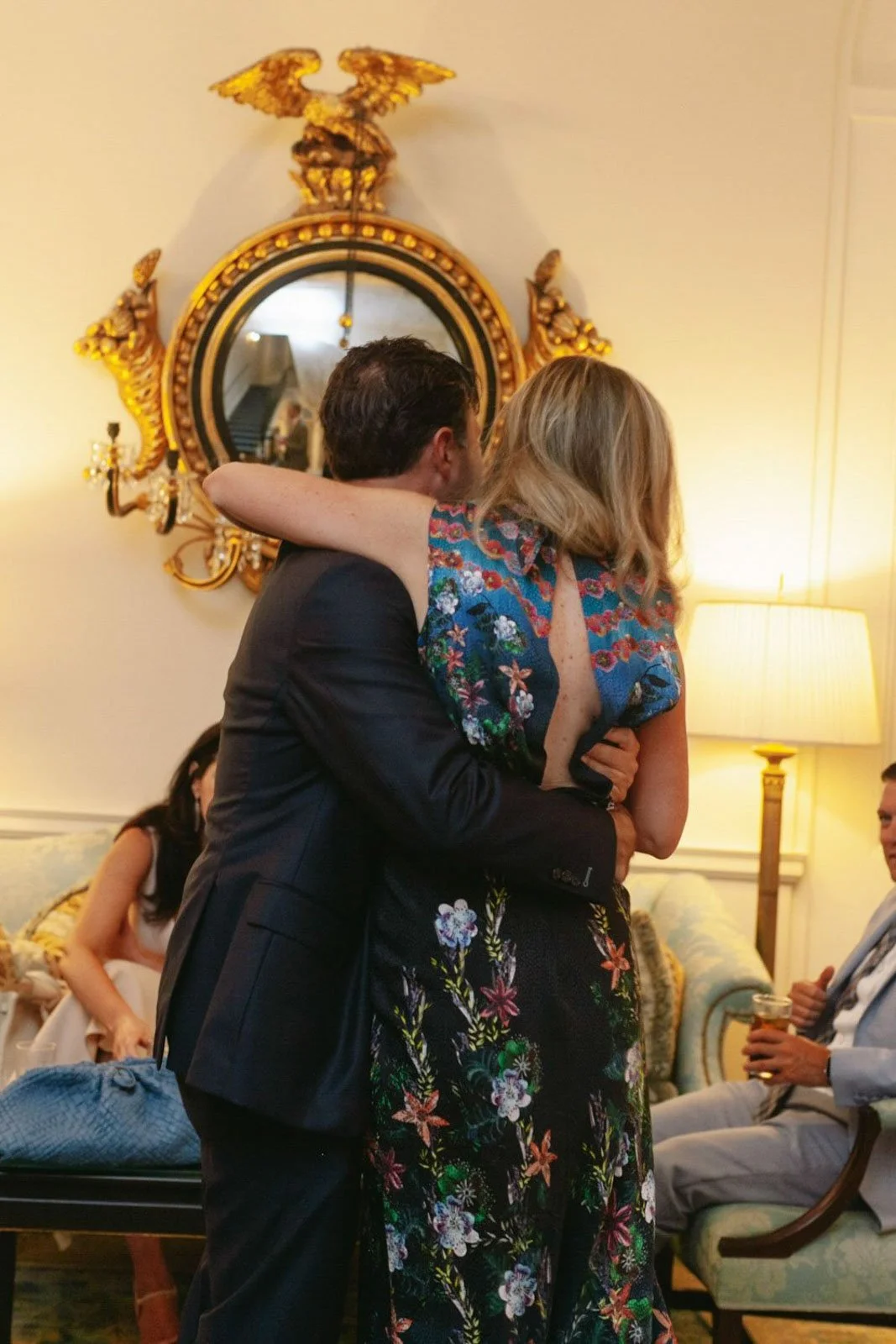 A man and woman hugging at a social gathering in a well-lit room with ornate decor, including a mirror with gold embellishments and a lamp.