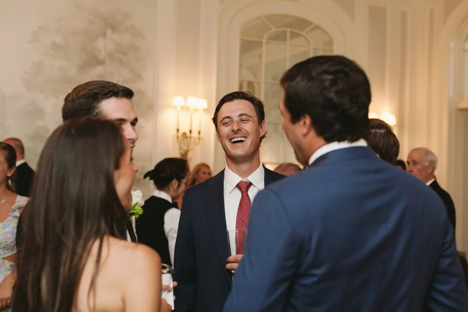 A group of people in formal attire socializing at an indoor event, with a man in a suit and tie laughing in the center.