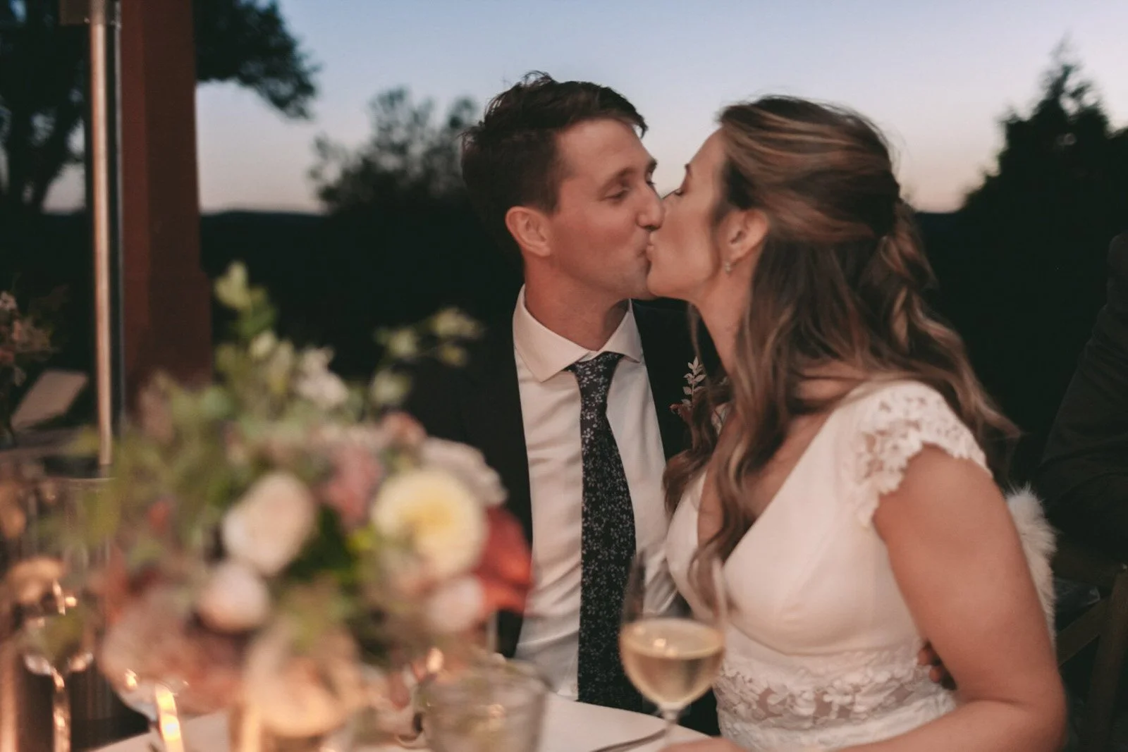 A couple sharing a kiss at a wedding reception, with flowers and a glass of white wine on the table in front of them, during evening time outdoors.
