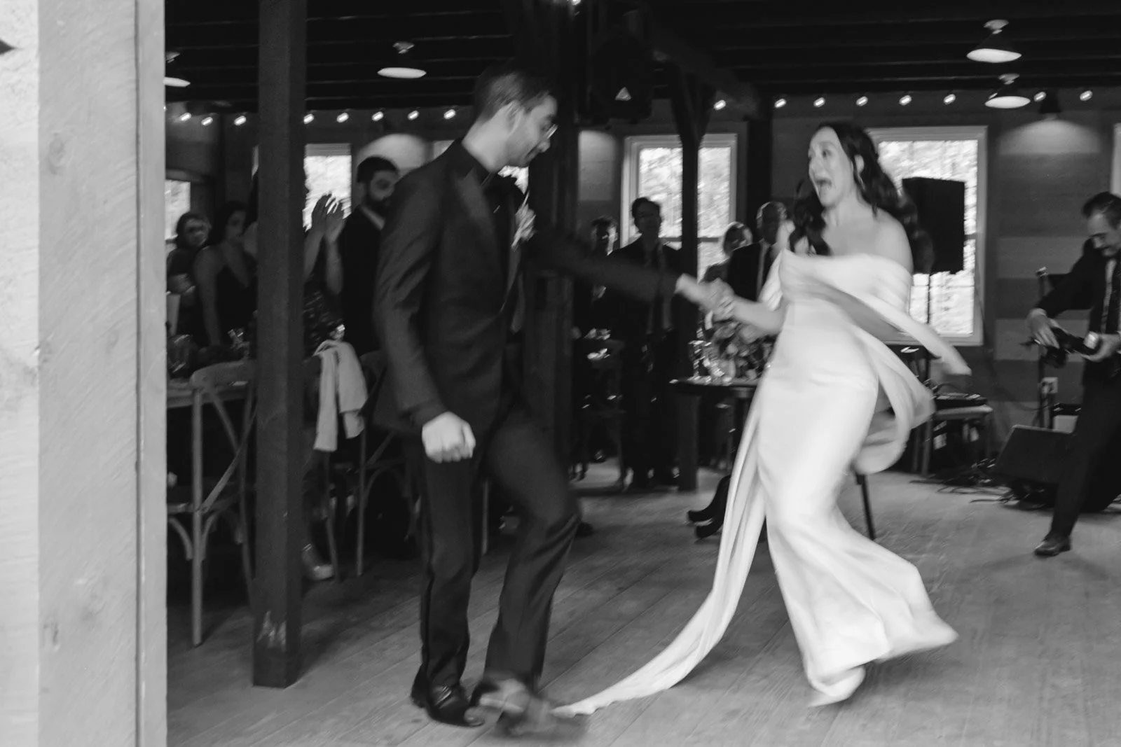 A bride and groom dancing at a wedding reception, surrounded by guests in a rustic indoor setting.