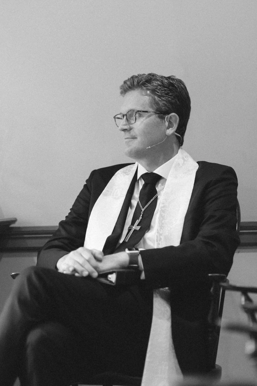A man wearing glasses, a suit, and a clerical stole with a cross necklace, sitting with his hands clasped, in a contemplative pose, in black and white.