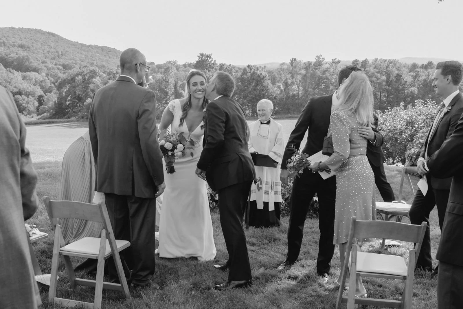 A black and white photo of a wedding ceremony outdoors with couples exchanging kisses and embraces, officiant in background, chairs, and scenic landscape with trees and hills.
