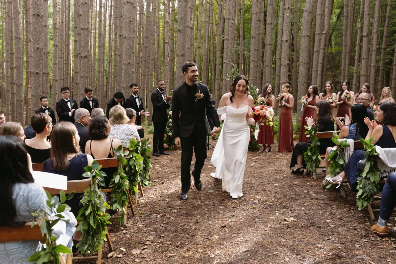 A couple walks hand-in-hand down the aisle during their outdoor wedding ceremony in a forest, surrounded by guests seated on wooden chairs decorated with greenery. The bride wears a white dress and holds a bouquet, while the groom is in a black suit.