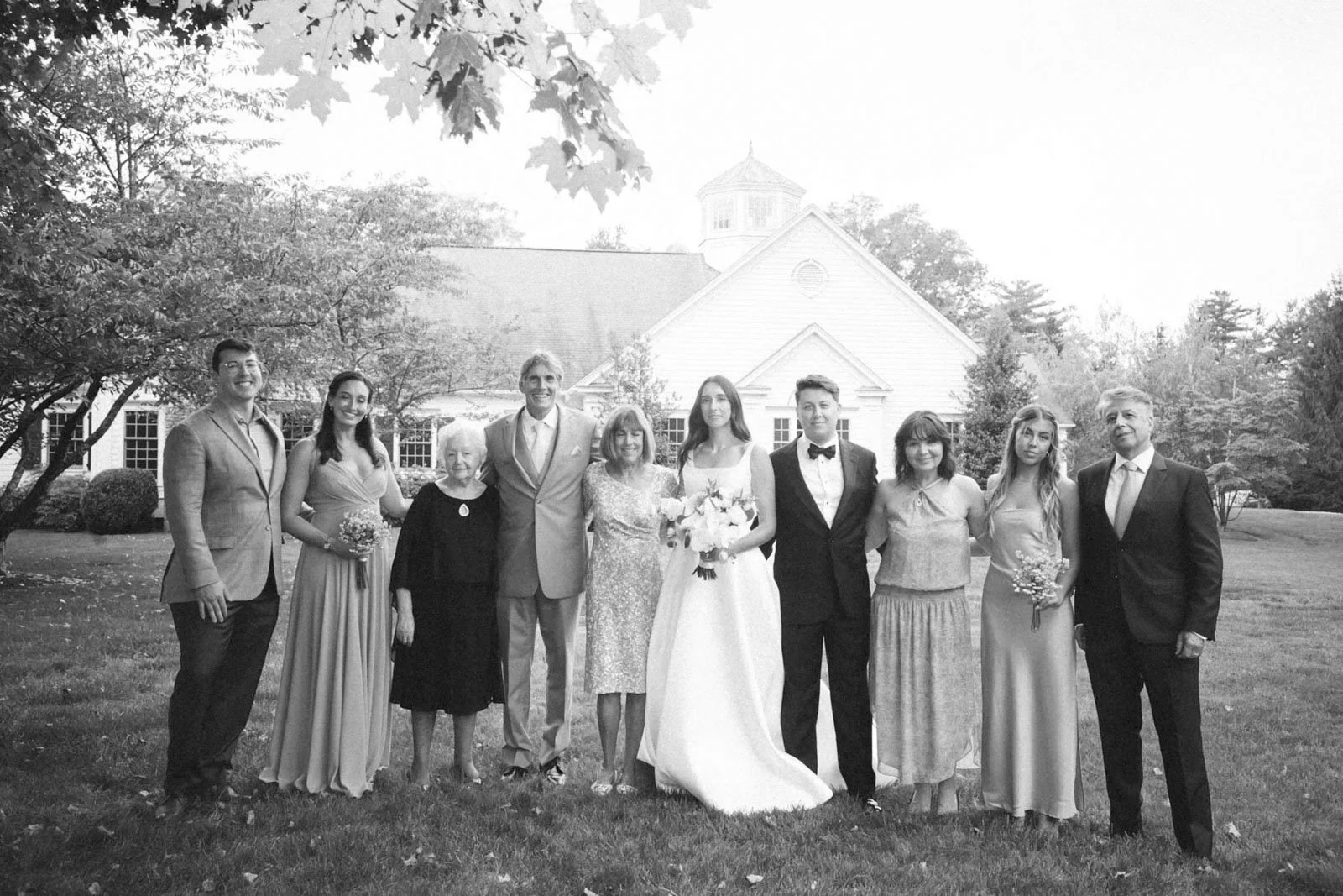 A black and white photo of a wedding group standing outdoors on grass in front of a white church building with trees in the background. The group includes the bride and groom, along with family and friends, all dressed in formal attire.