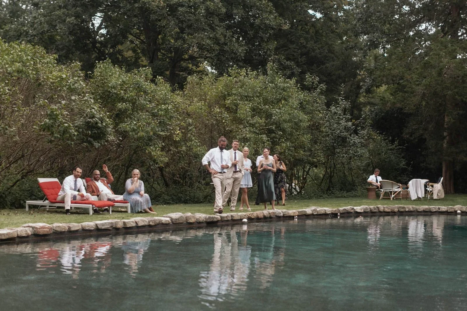 People relaxing and socializing by a pond in a backyard with outdoor furniture and lush green trees.