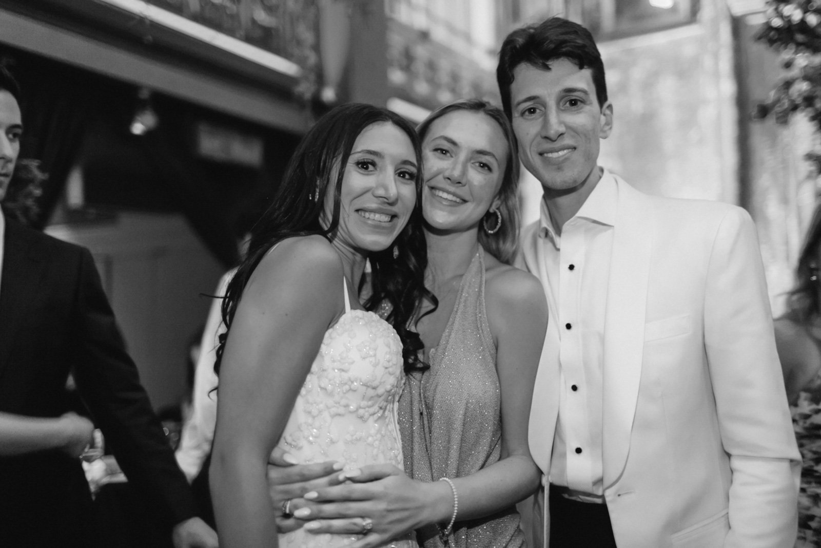 Three people smiling on a city street in black and white, two women and one man dressed in formal attire.