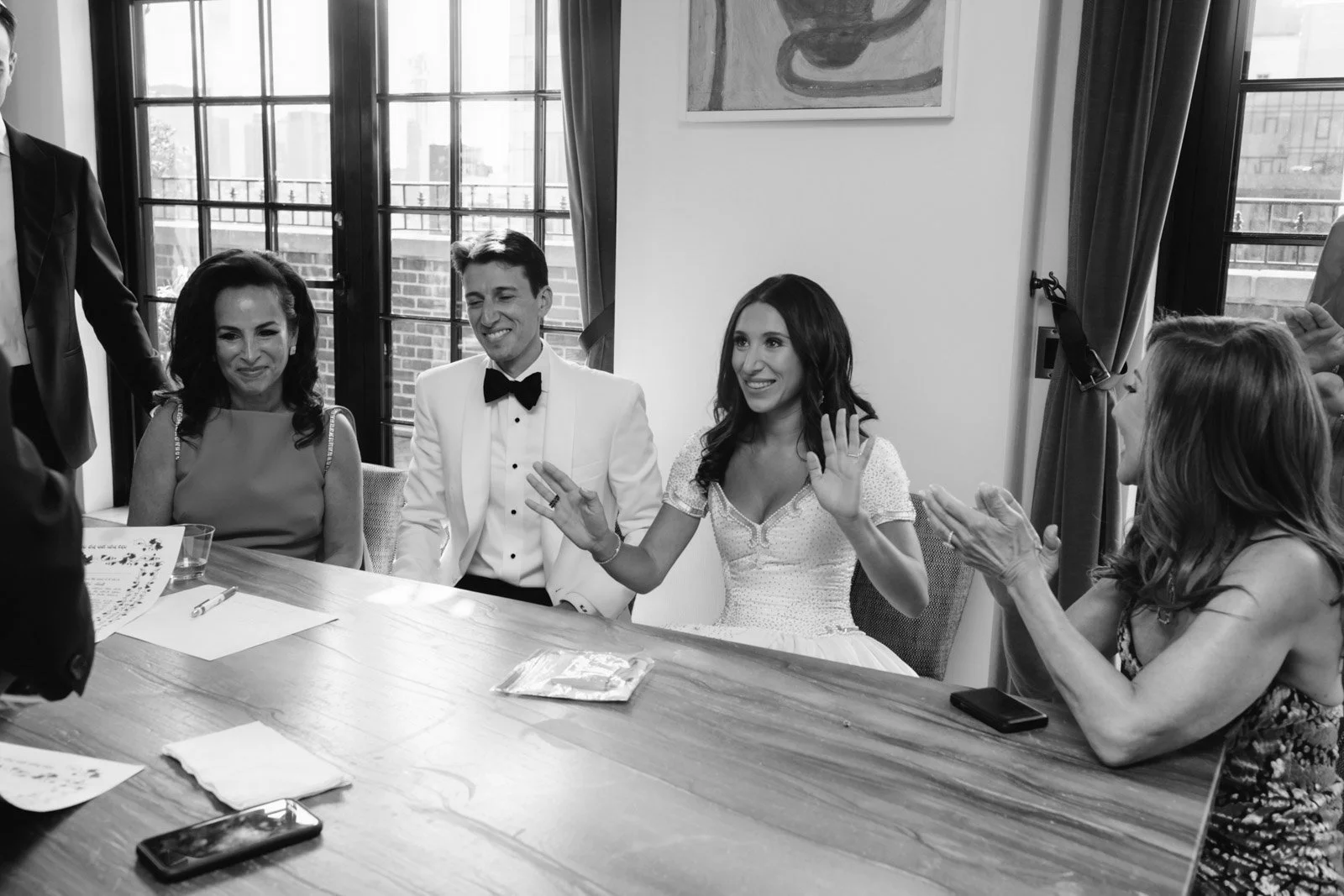 A group of five people sitting around a table, celebrating a wedding ceremony. The bride and groom are smiling and showing their wedding rings, while the other guests are clapping and smiling.