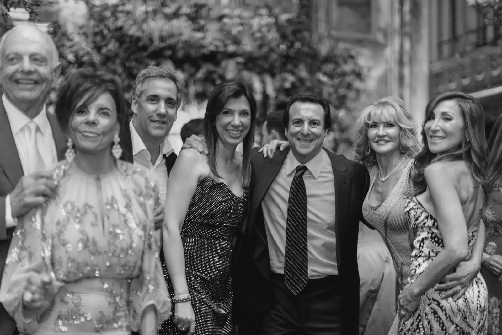 A group of seven people, four women and three men, smiling and posing together at a social event, with a tree and building in the background.