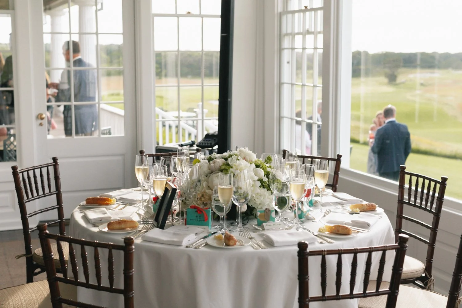 A round table set for a formal event with white tablecloth, floral centerpiece, champagne glasses, and place settings, inside a bright room with large windows overlooking a grassy outdoor area where people are gathered.
