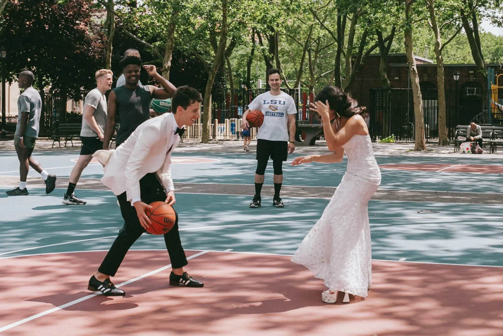 A wedding couple is playing basketball on an outdoor court surrounded by trees, with friends watching and laughing.