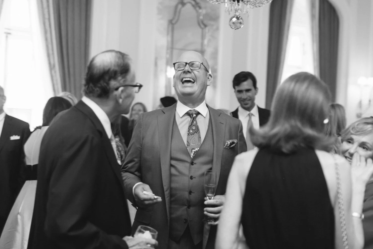 People at a social gathering, laughing and chatting, in a formal setting with elegant decor.