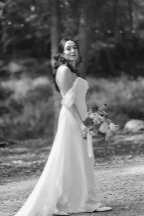 A woman in a flowing dress standing outdoors holding a bouquet of flowers, with trees and nature in the background.
