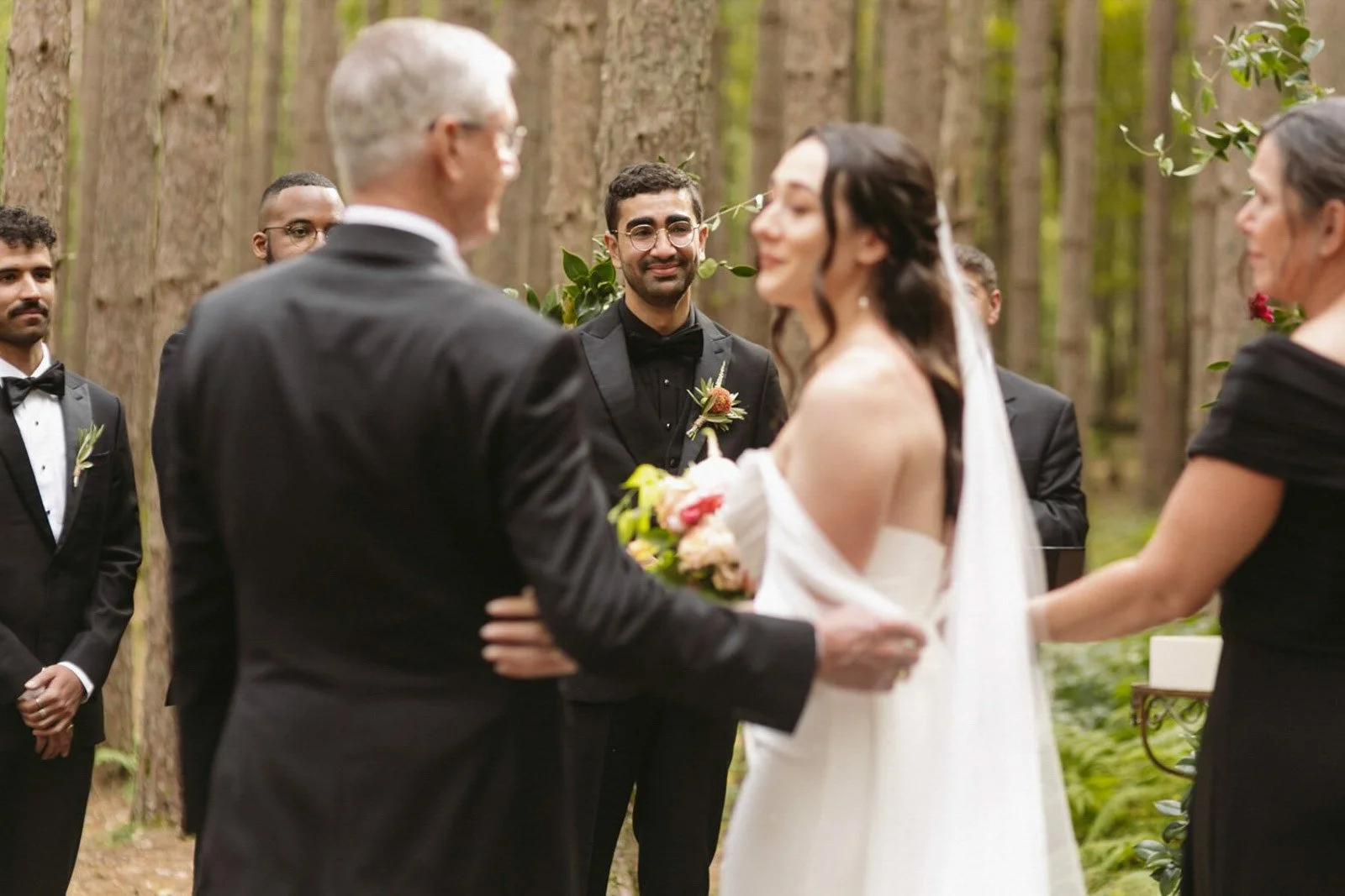 A wedding ceremony takes place in a forest with tall trees. The bride, in a white off-shoulder dress, holds a bouquet of flowers and faces the groom, an older man in a black suit. The groom, in a black suit and bow tie, stands across from her with an