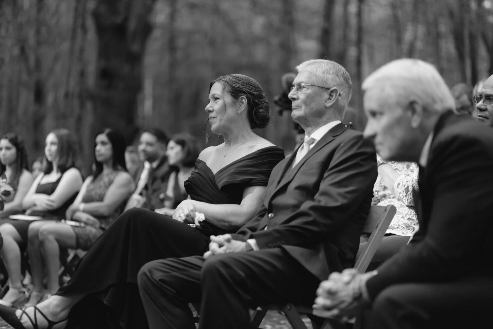 People attending an outdoor formal event, sitting in chairs in a wooded area, dressed in formal attire.