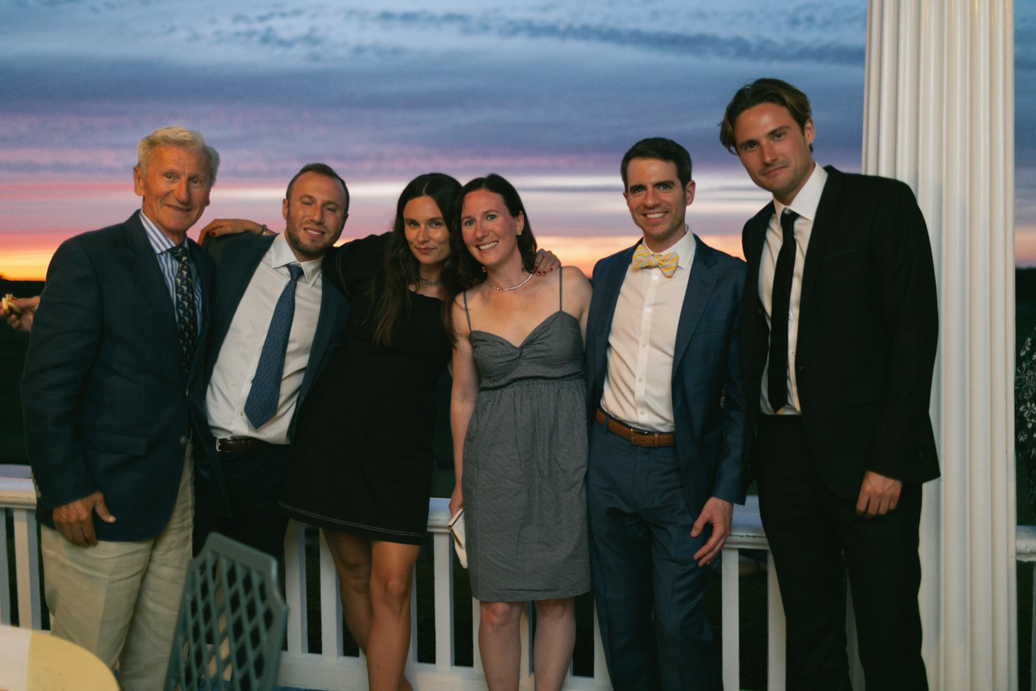 Group of six people dressed in formal attire at sunset on an outdoor porch or balcony.