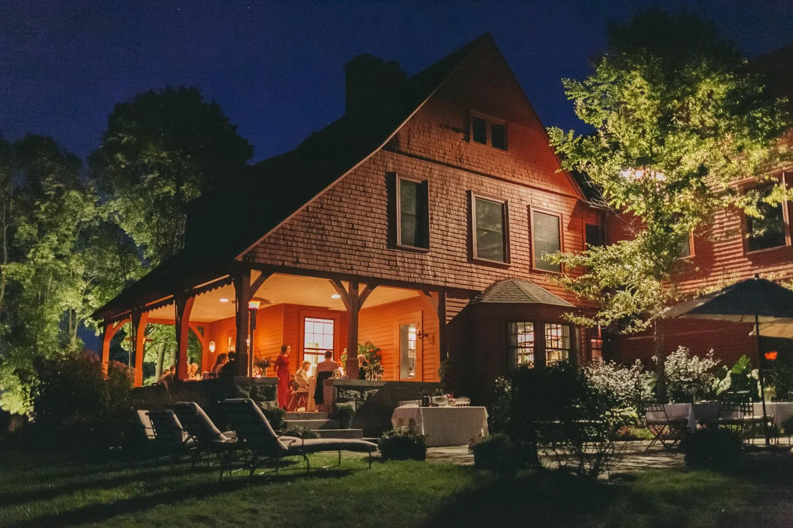 Nighttime outdoor gathering on a covered porch of a large, red, wooden house surrounded by trees and furniture.