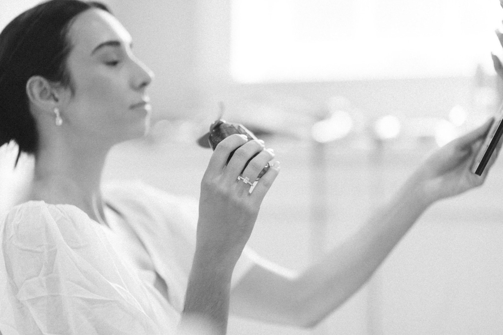 A woman with dark hair wearing a white top is holding a phone and a bottle, with her eyes closed in a relaxed pose.