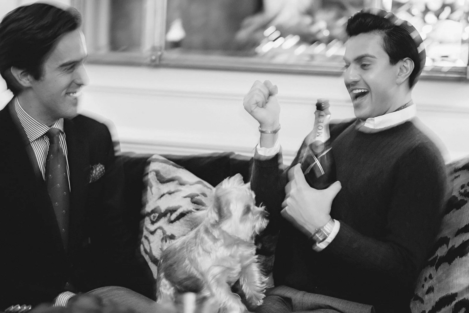 Two men are smiling and laughing while sitting on a couch next to a small dog. One man is holding a champagne bottle. The scene appears to be cheerful and relaxed.