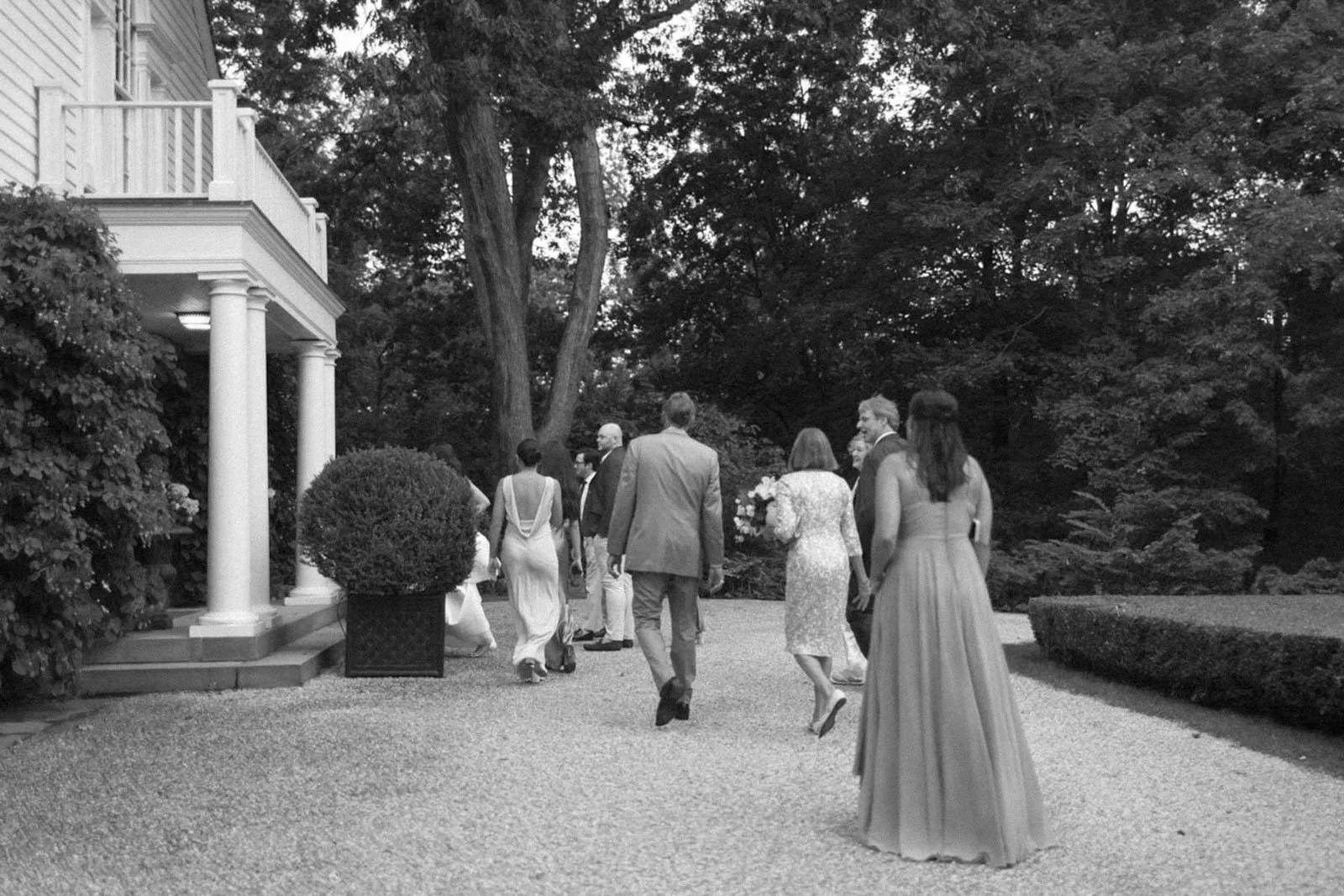 A group of people dressed in formal attire walking outside on a gravel path near a house with columns and surrounded by trees and bushes, in black and white.