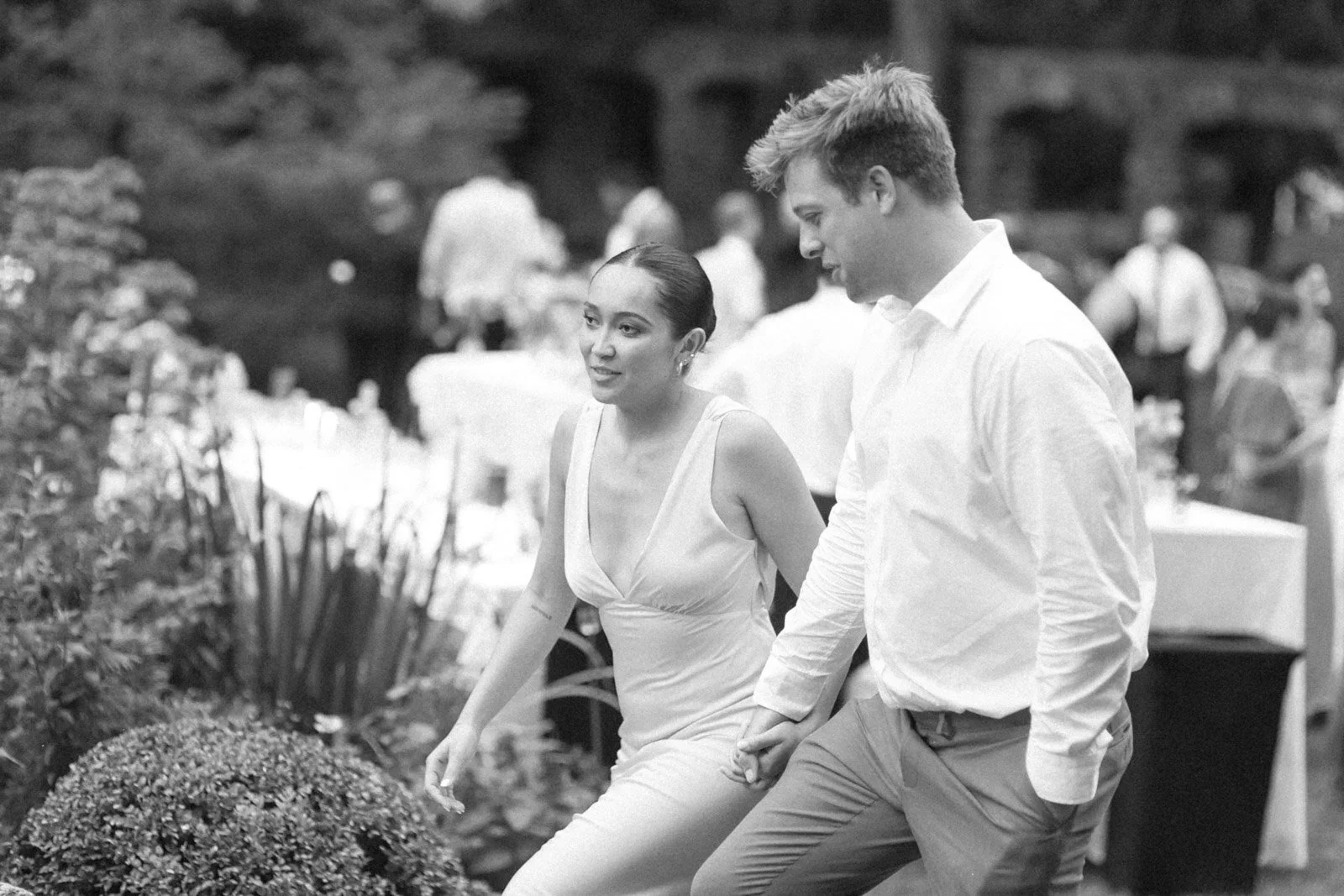A black and white photo of a woman and a man holding hands and walking outdoors at an event with booths or tables in the background.