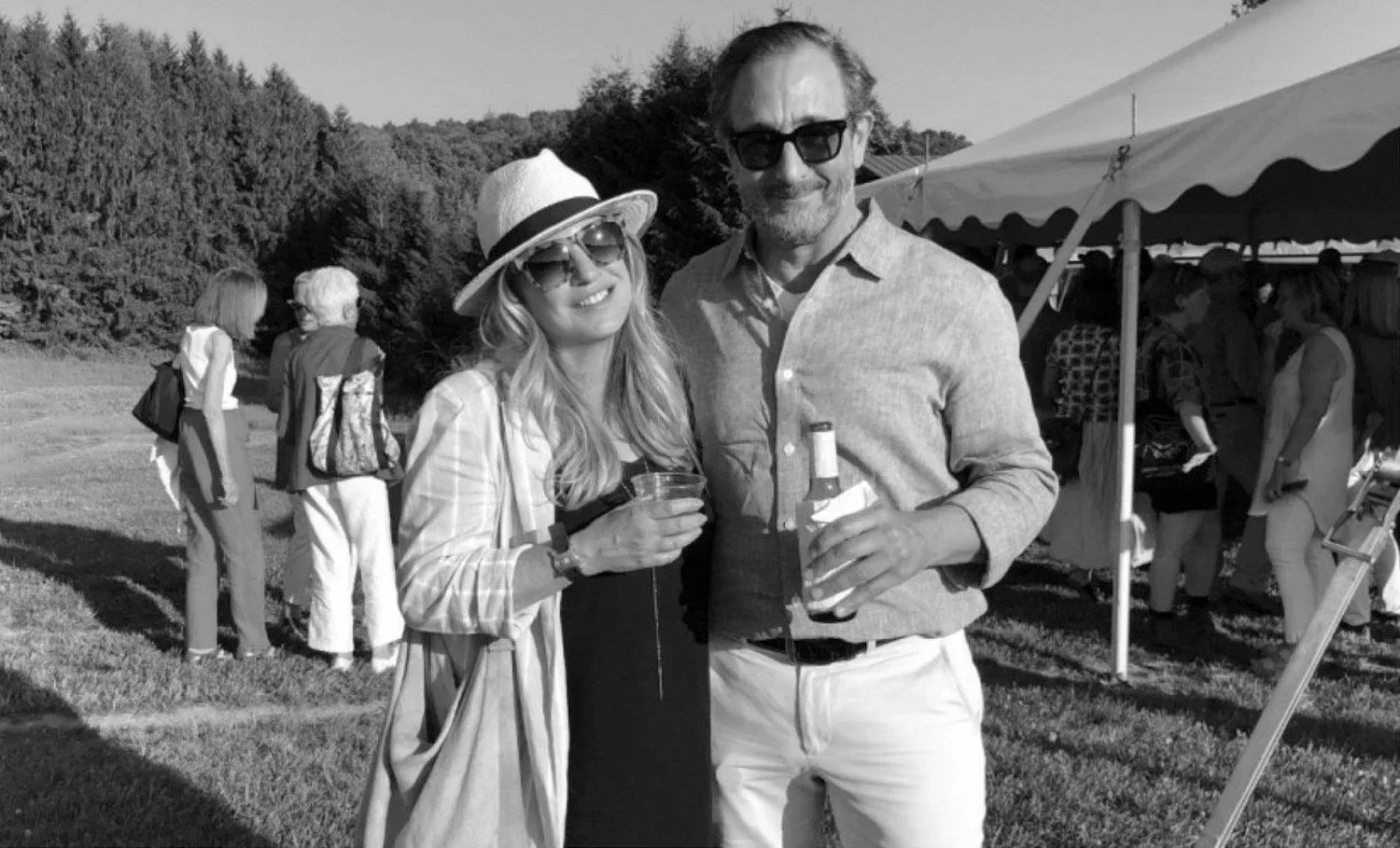 A black-and-white photo of a man and woman smiling and holding drinks at an outdoor event with a tent and people in the background.