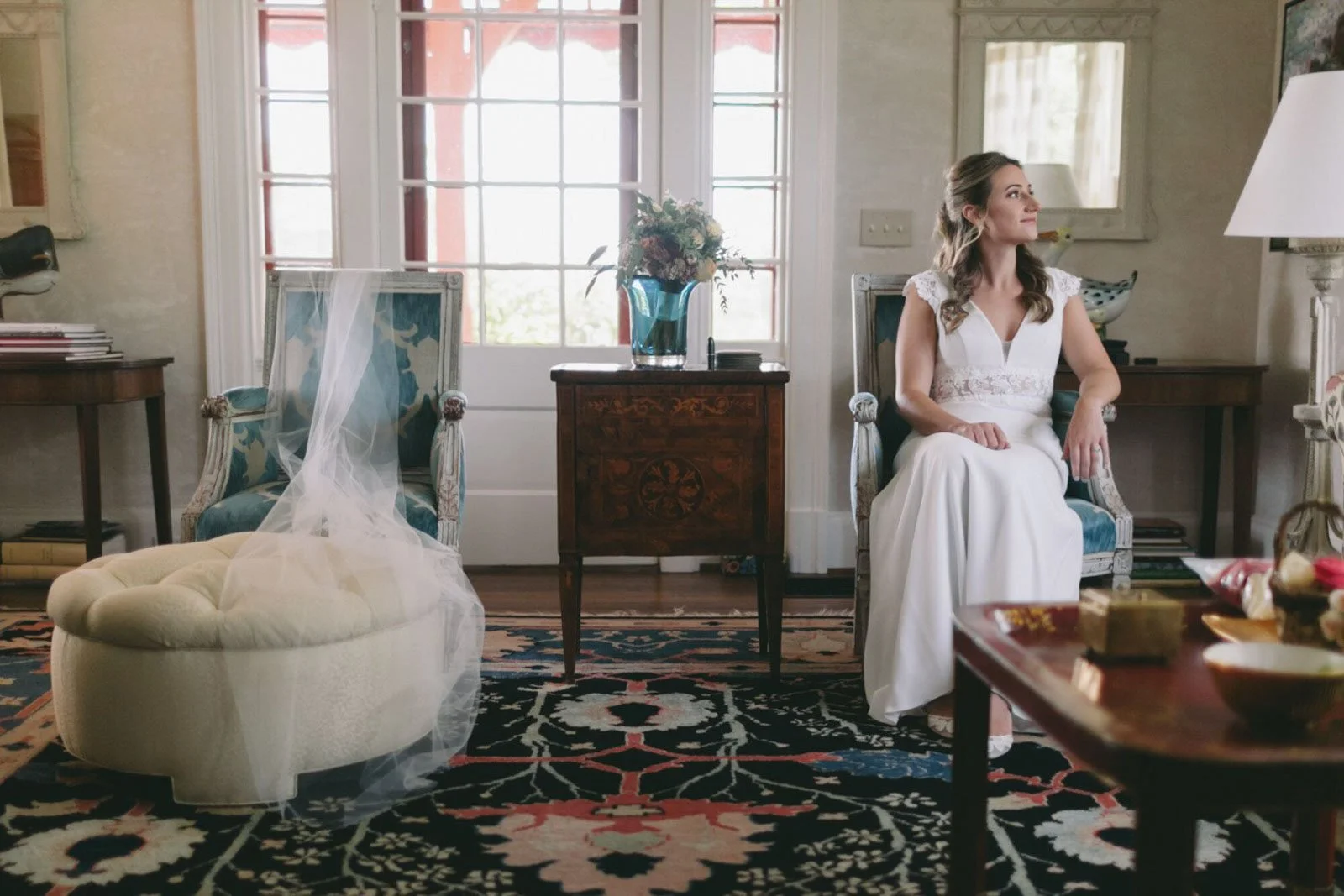A woman in a white dress sitting on a vintage armchair in a well-lit room with a large window, patterned rug, and various decorative items, possibly preparing for a wedding.