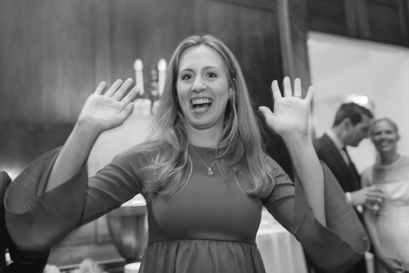 A woman with long hair smiling and raising both hands, in an indoor setting with other people in the background.