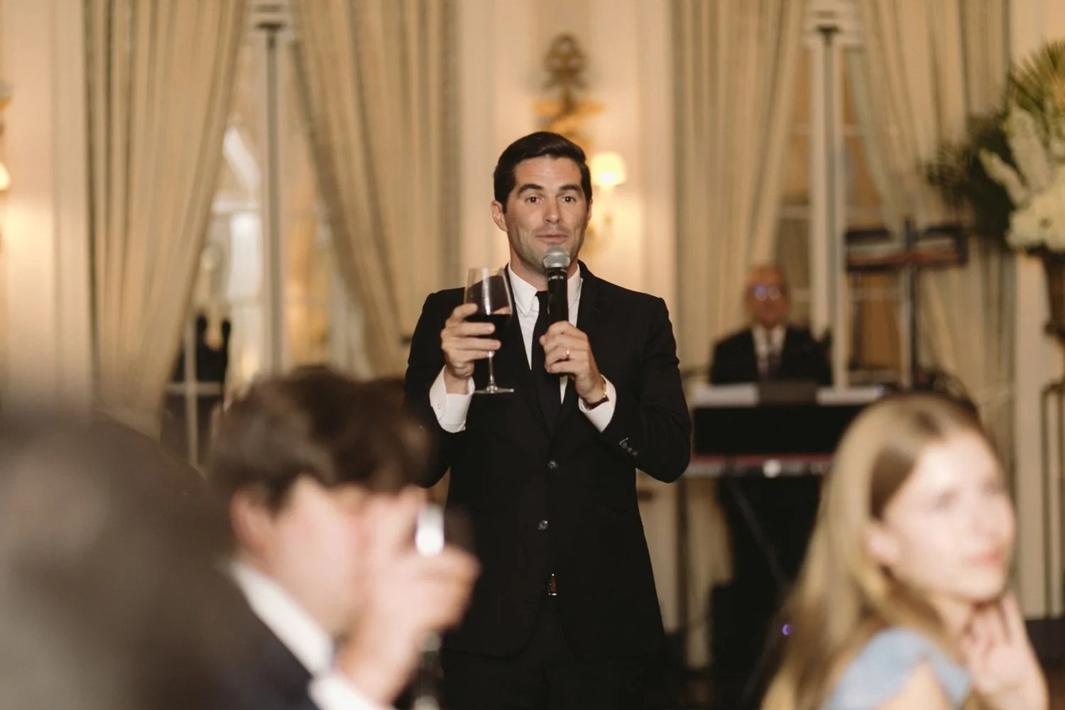 A man in a black tuxedo holding a glass of red wine and speaking into a microphone at a formal event or wedding reception.