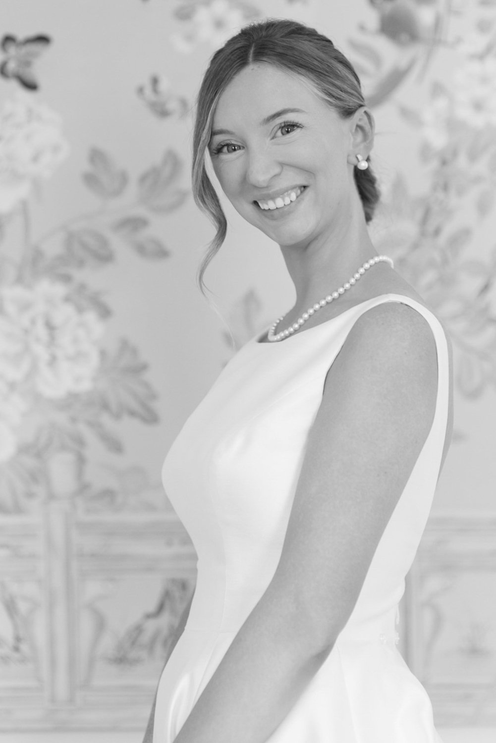 A smiling woman with shoulder-length hair, wearing a sleeveless white dress, pearl necklace, and pearl earrings, standing in front of a painted floral background.