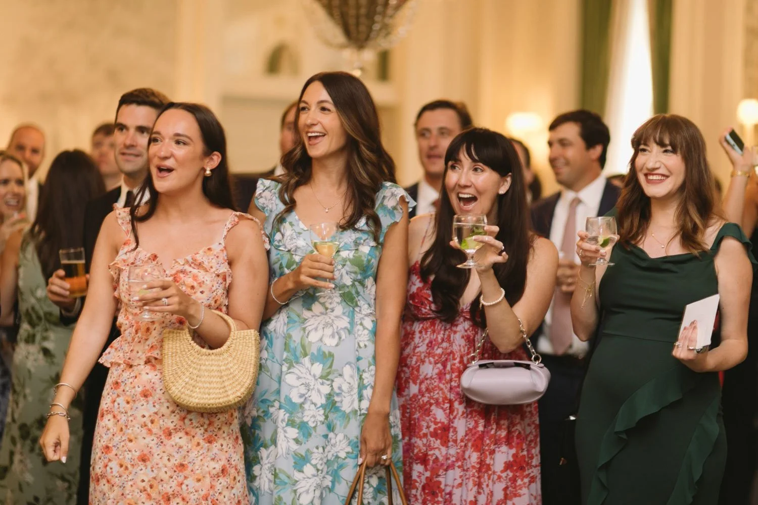 Group of women and men at a social event, smiling and holding drinks, dressed in formal and semi-formal attire.