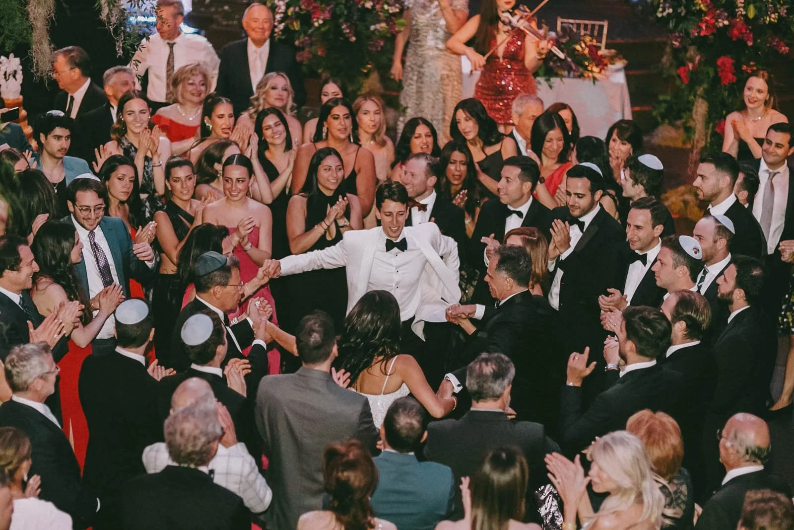 A wedding celebration with the bride and groom dancing in the center surrounded by guests clapping and cheering. The bride is in a white dress and the groom in a white tuxedo, all set in an elegant decorated venue with flowers.