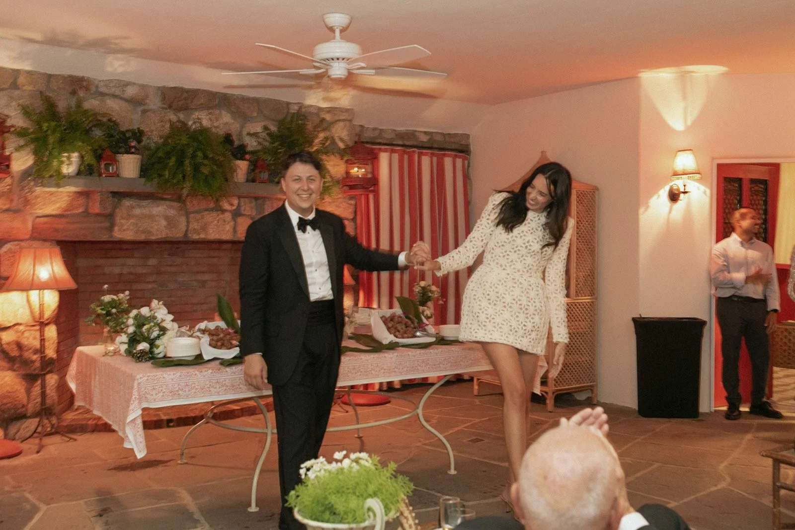 A couple dressed in formal attire, holding hands and smiling at a celebration or event in a warmly decorated room.