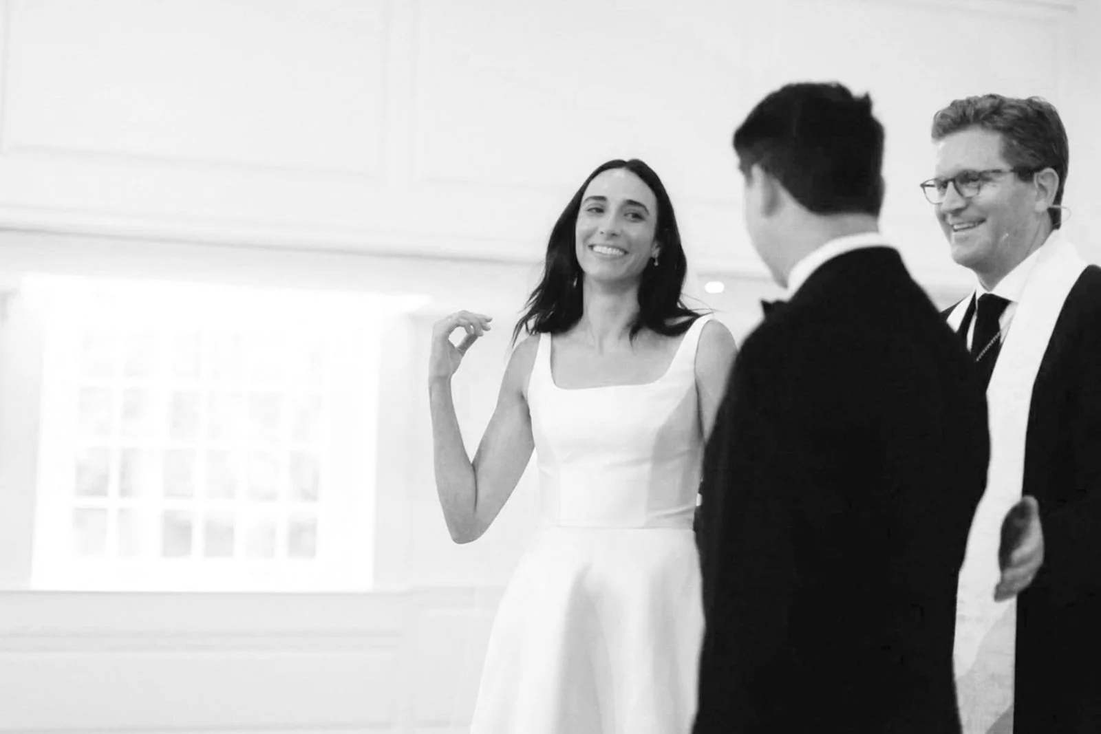 A woman in a white dress smiling and talking to two men in tuxedos at a formal event or wedding, in black and white.