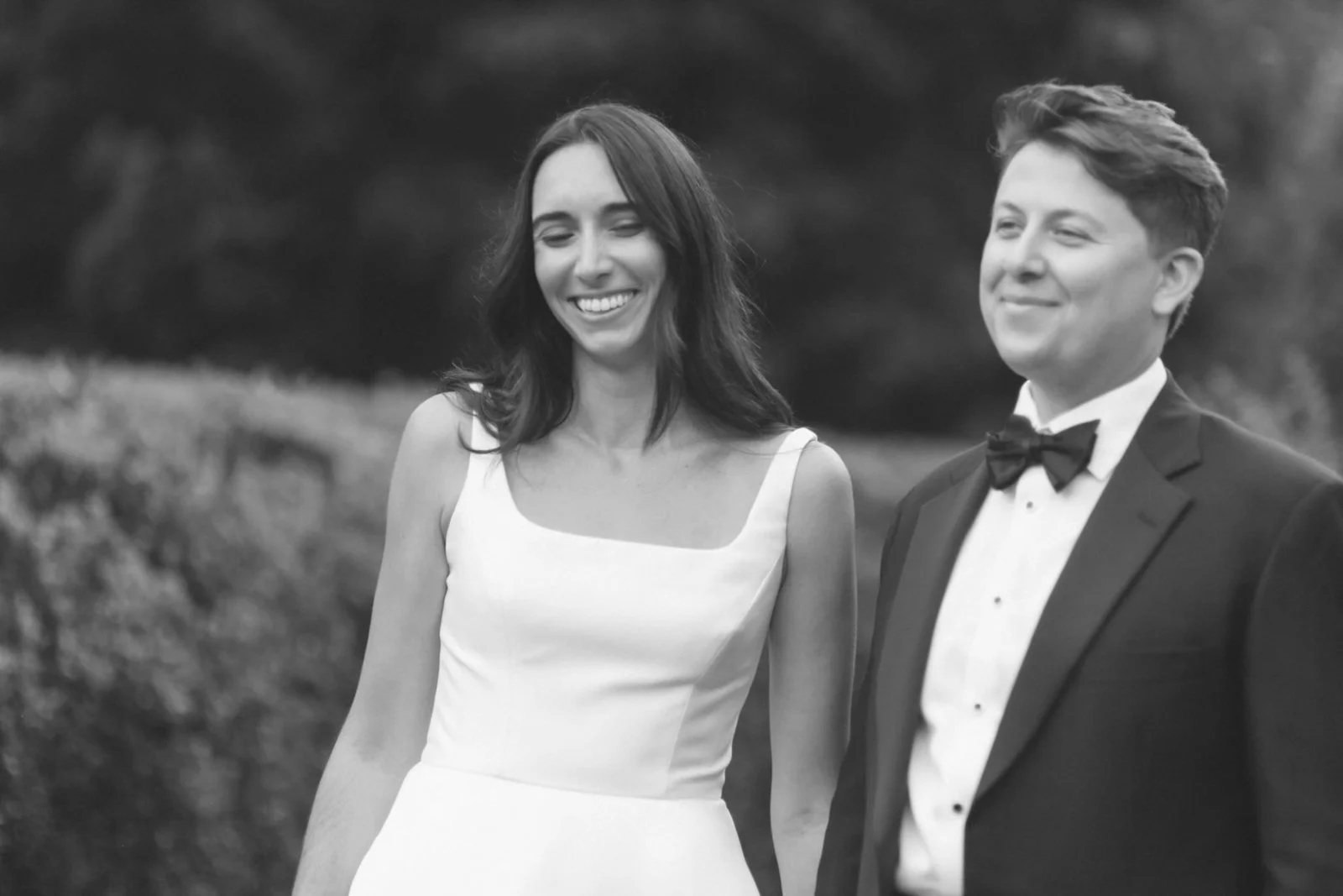 A black and white photo of a smiling woman in a white dress and a man in a tuxedo with a bow tie, standing outdoors with a blurred natural background.