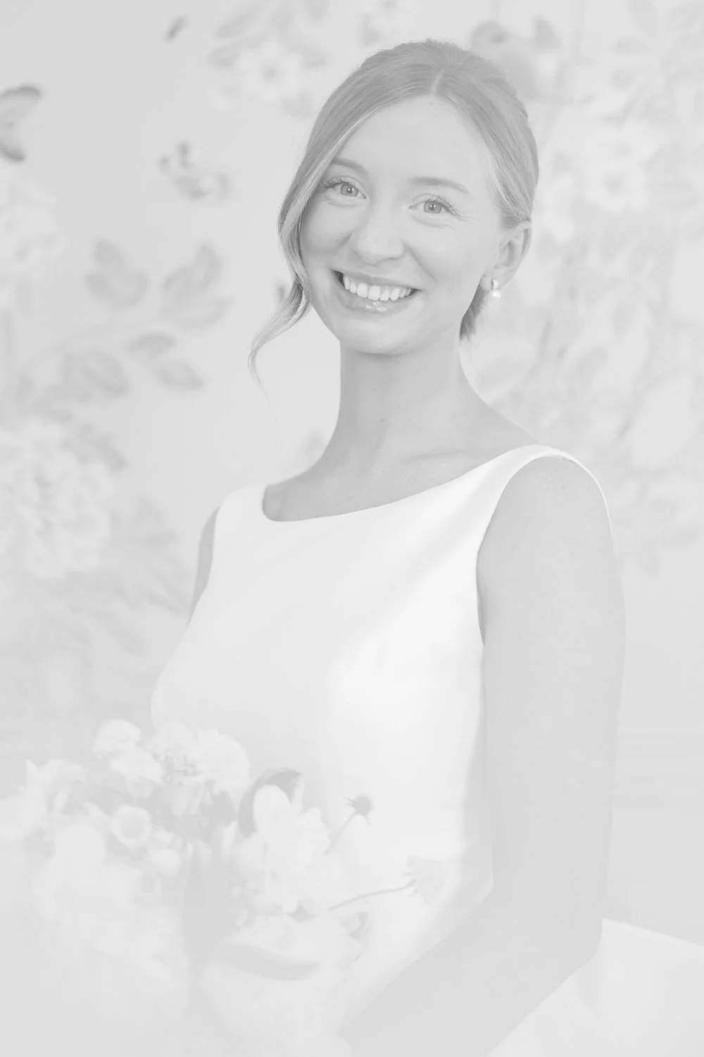Portrait of a smiling woman in a white dress, holding a bouquet of flowers, standing in front of a patterned background.